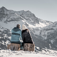 <p>Paar sitzt auf Holzstämmen im Schnee und schaut auf das schöne winterliche Bergpanorama.</p>