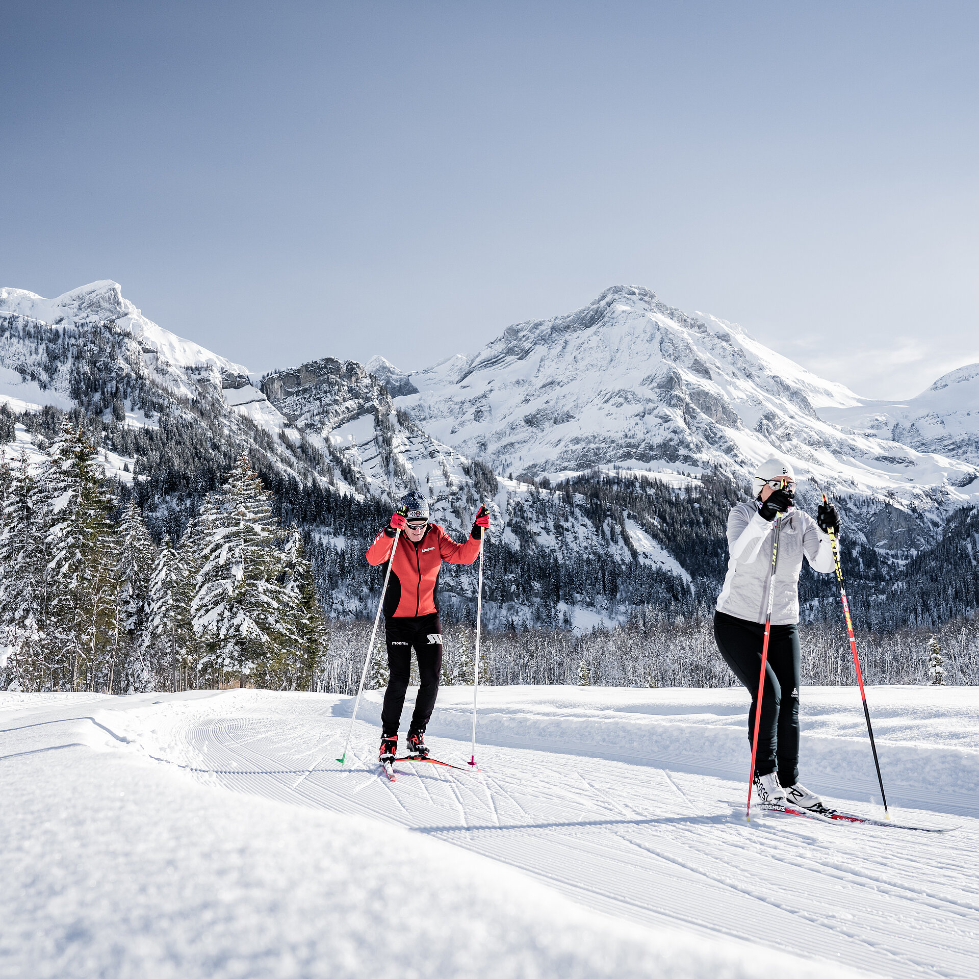 <p>Ein Ehepaar, in rot und weiss gekleidet, am Langlaufen auf einer Loipe vor verschneitem Bergpanorama.</p>