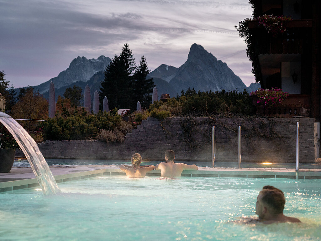 <p>Drei Personen schwimmen in einem Whirlpool im Freien, mit Bergen im Hintergrund.</p>