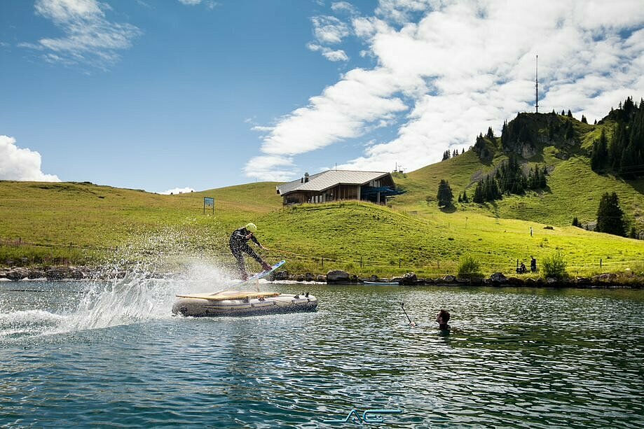 <p>Eine Person auf einem Wakeboard fährt auf einem kleinen Bergsee über verschiedene Schanzen.</p>