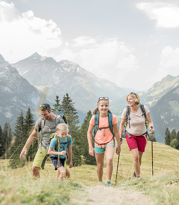 <p>Eine Familie wandert auf einem Wanderweg neben einer grünen Wiese, im Hintergrund eine Berglandschaft.</p>