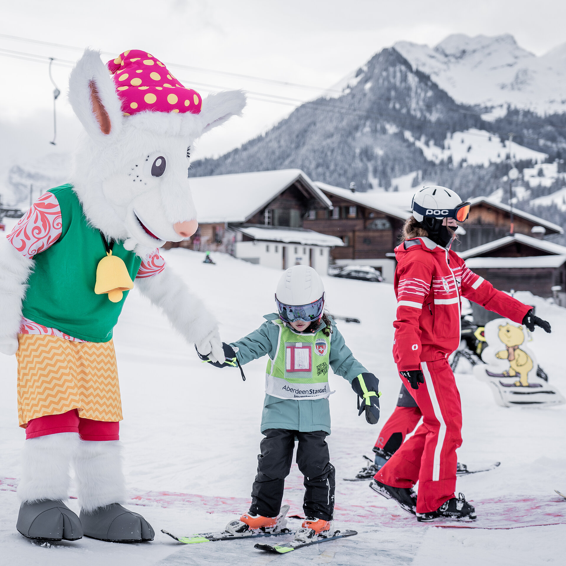 <p>Das Maskottchen Saani im Kinderland der Skischule Gstaad mit einer rot gekleideten Skilehrerin und zwei kleinen Kindern auf Ski.</p>