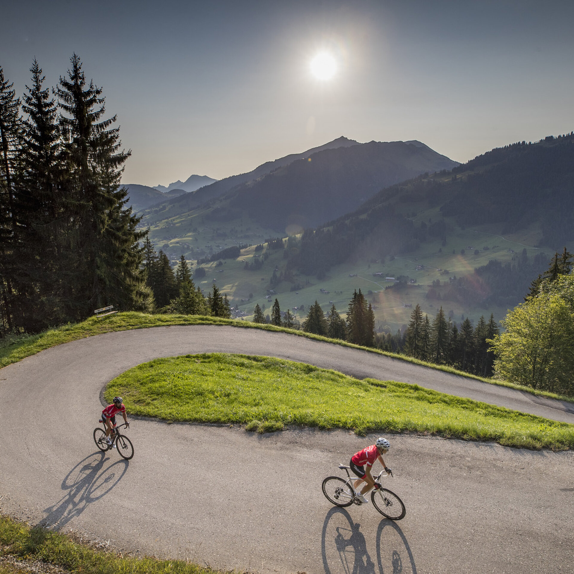 <p>Zwei Rennradfahrer (eine Frau und eine Mann) fahren in einer steilen Kehre bergauf. Am Himmel sieht man die Sonne in einem milchig-blauen Himmel. Es ist Sommer.</p>