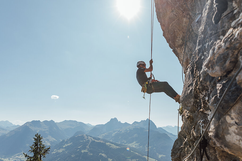 <p>Ein Kletterer seilt sich an einem Seil den Fels hinab, im Hintergrund die Bergkulisse vom Saanenland unter blauem Himmel.</p>