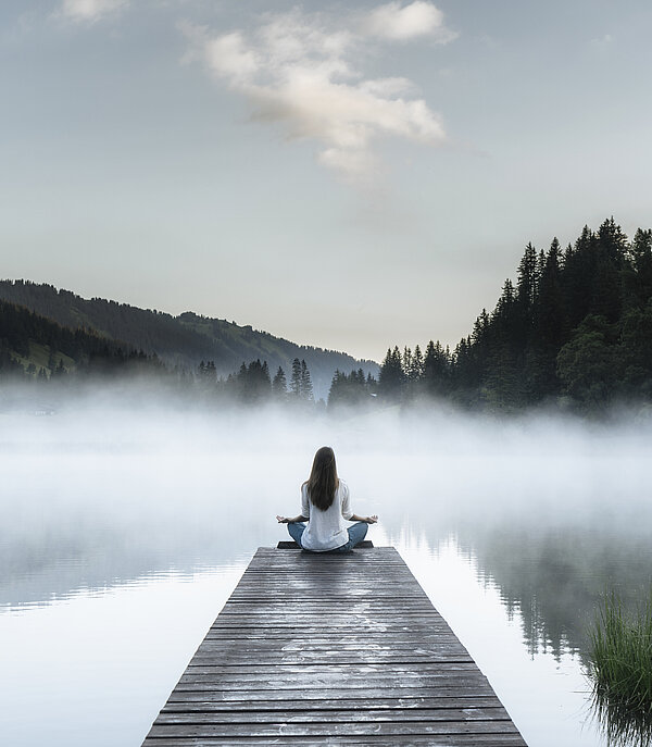 <p>Frau macht Entspannungsübungen auf einem Steg beim See, welcher leicht im Nebel eingehüllt ist.</p>