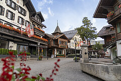 Promenade with houses decorated with flowers, trees, red flowers in the foreground, a fountain on the right. Flags with Swiss, Bernese and Saanen coats of arms and Gstaad Menuhin Festival.