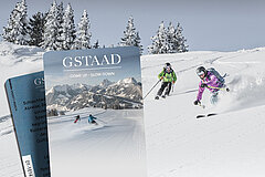 Two skiers descending through powdery snow, with snow-covered fir trees in the background. In the foreground are two ski keycards with the Gstaad logo and a picture of a ski slope and two skiers against a panoramic mountain backdrop.