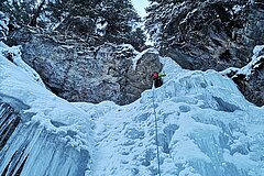 Person klettert gesichert eine vereiste Felswand hinauf, umgeben von Schnee, Eis und winterlichem Wald.
