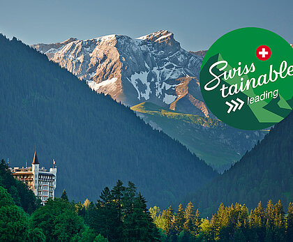 Links im Vordergrund des Bildes sieht man das Hotel Palace - ein weisses Schlossgebäude. Darum herum sieht man viele grüne Bäume, bewaldete Hügel und zu hinterst im Bild Berge mit etwas Schnee drauf. Auf der rechten oberen Seite ist ein grüner Kleber positioniert. Darauf steht Swisstainable III - Leading und eine kleines Schweizerkreuz.