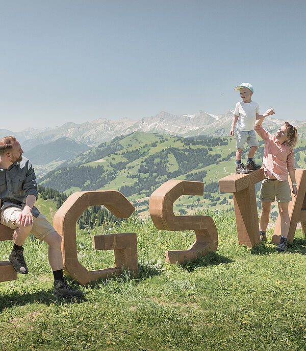 Family of four standing and sitting on the wooden logo «Gstaad» Rinderberg in summer.