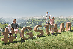 Eine vierköpfige Familie steht und sitzt auf dem Holz-Logo «Gstaad» Rinderberg im Sommer.