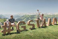 Eine vierköpfige Familie steht und sitzt auf dem Holz-Logo «Gstaad» Rinderberg im Sommer.