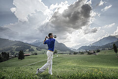 Un golfeur en pantalon blanc et chemise bleue, joue au golf devant un panorama de montagnes.