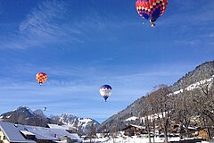 Mehrere bunte Heissluftballone über verschneiten Häusern und Bergen im klaren Winterhimmel.