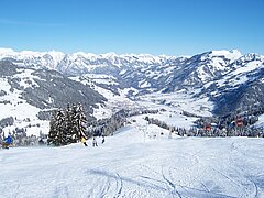  Piste de ski photographiée en vue aérienne dans la direction de la vallée. Zweisimmen visible en arrière-plan, au milieu des montagnes enneigées. 
