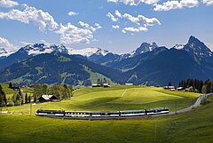 Devant on voit une composition de train bleu foncé/blanc, derrière laquelle on voit des collines vertes et, tout au fond, des sommets de montagne enneigés.