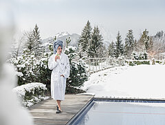 Une femme vêtue d’un peignoir blanc, les cheveux enveloppés dans une serviette bleue, est installée au bord d’une piscine extérieure avec un verre dans sa main droite. En arrière-plan, la forêt et les montagnes recouvertes de neige.