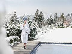 Une femme vêtue d’un peignoir blanc, les cheveux enveloppés dans une serviette bleue, est installée au bord d’une piscine extérieure avec un verre dans sa main droite. En arrière-plan, la forêt et les montagnes recouvertes de neige.