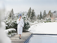 A woman wearing a white bathrobe and with a blue towel wrapped around her head, holding a glass in her right hand and standing next to an outdoor swimming pool, with snow-covered forest and mountains in the background.