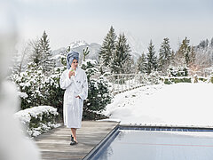 Eine Frau in weissem Bademantel und mit blauem Tuch um den Kopf mit einem Glas in der rechten Hand steht neben einem Aussenschwimmbecken. Im Hintergrund verschneiter Wald und Berge.