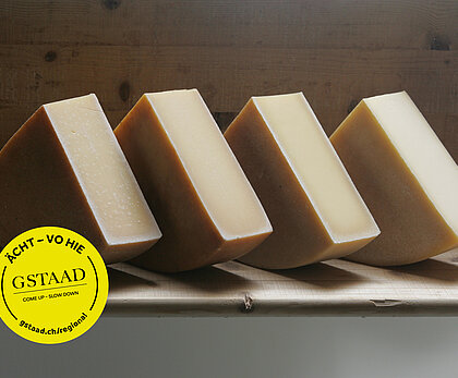 Auf einem Holzbrett stehen viermal ein Viertel eines Hartkäses. Die Wand dahinter ist auch aus Holz. Auf der linken unteren Seite ist ein gelber, runder Kleber platziert. Darauf steht Gstaad - Come up - Slow down in der Mitte und am Rand jeweils ÄCHT - VO HIE und gstaad.ch/regional