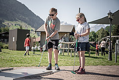 Zwei Kinder stehen auf einem Minigolf Platz und versuchen den Ball ins Loch zu schiessen.