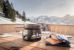 Schwarzes Fonduecaquelon und Brot stehen im überdimensionalen Fonduecaquelon aus Holz bereit, Aussicht auf winterliche Landschaft mit Bäumen und Bergen.