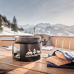 Black fondue caquelon and bread in a giant wooden fondue caquelon, against a wintry backdrop of trees and mountains.
