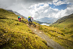 Gruppe von Bikern auf einem Bergtrail bei Sonnenuntergang, mit weitem Blick auf Berge und Wolken.