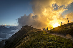Gruppe von Bikern auf einem Bergtrail bei Sonnenuntergang, mit weitem Blick auf Berge und Wolken.