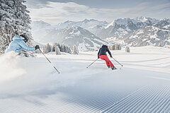 Couple skiing down a freshly prepared ski slope, with snow-covered trees and mountains in the background.