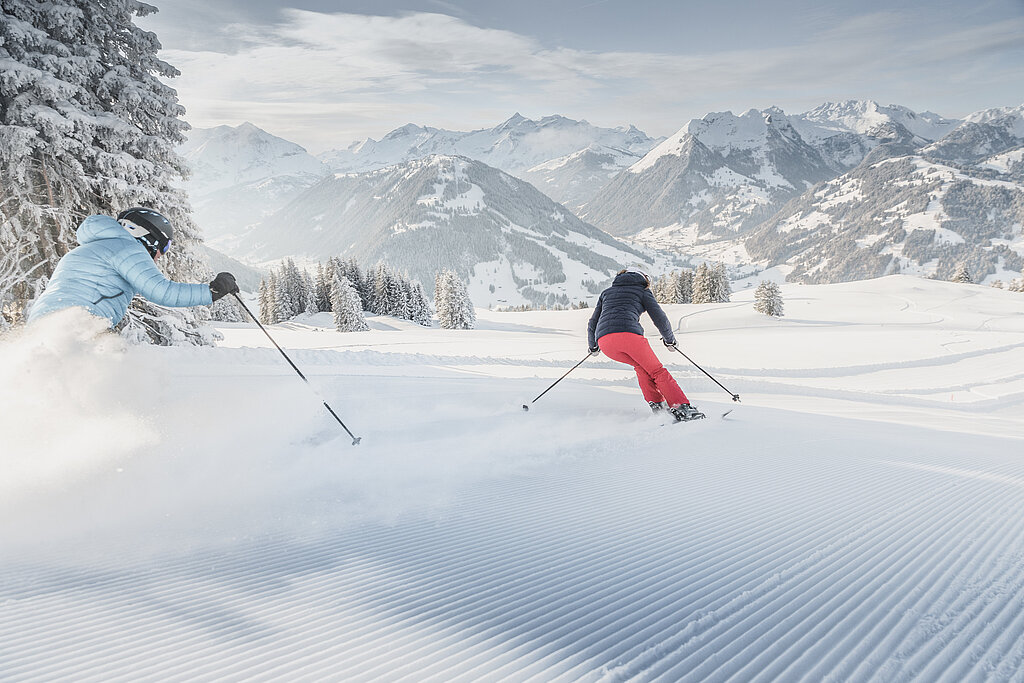 <p>Ein Paar fährt mit Skiern die frisch präparierte Skipiste hinunter. Im Hintergrund schneebedeckte Bäume und Berge.</p>