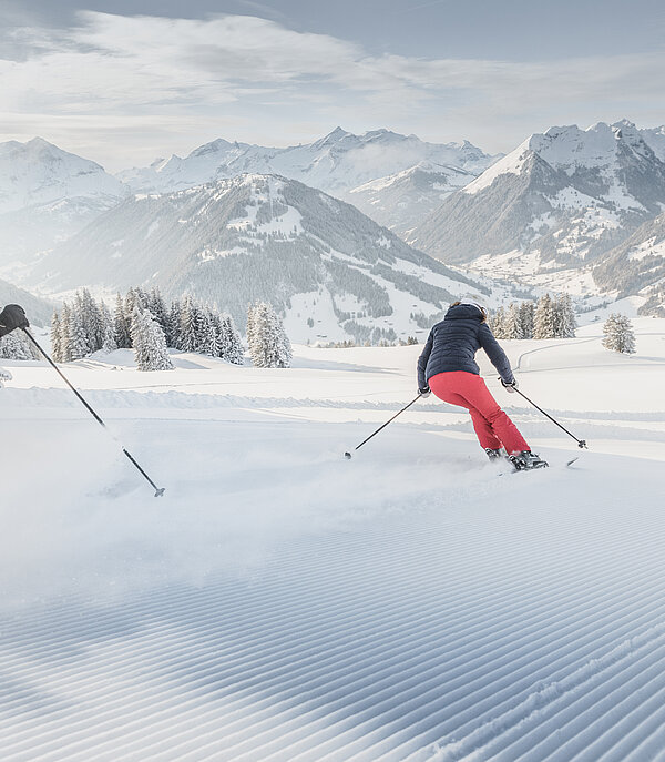 Un couple descend la piste fraîchement damée à skis. En arrière-plan, des arbres et des montagnes recouverts de neige.