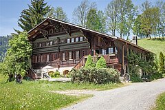 Altes Haus in typischer Saanen Chalet-Architektur umgeben von Bäumen und Grünfläche mit Kiesstrasse im Vordergrund.
