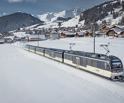 Eine dunkelblau/weisse Zugkomposition verlässt ein Dorf und fährt durch eine verschneite Landschaft. Der Zug besteht aus fünf Wagen.