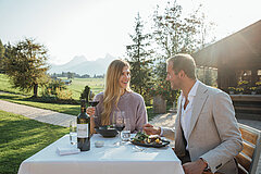 Couple sitting outdoors at a set table in front of a chalet, in the middle of mountain scenery.