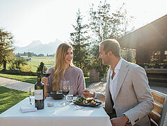 Pärchen sitzt im Freien an einem gedeckten Tisch, dahinter steht ein Haus, Bäume, Wiese und weiter fort schönes Bergpanorama.