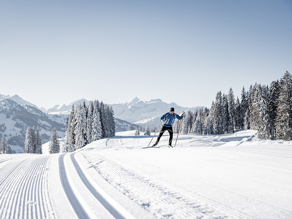 <p>Langlauf Sportler skatet über eine frisch gespurte Loipe, im Hintergrund befinden sich verschneite Berge</p>