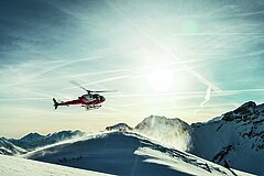 Helikopter landet auf schneebedecktem Berggrat, Schneewirbel im Sonnenlicht, umliegende Gipfel im Hintergrund.