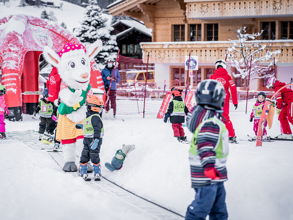 <p>Kleine Kinder in bunten Skianzügen auf Skis lassen sich zusammen mit Saani auf dem Rollband transportieren.</p>