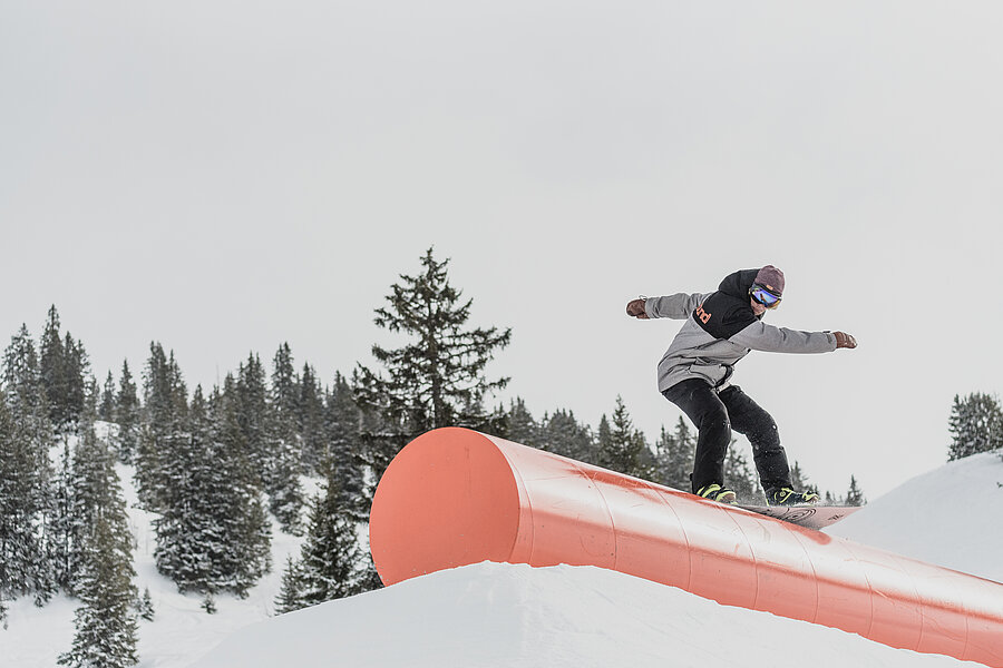 A dark-clad snowboarder rides over an orange rail in the snowpark.