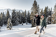 Ein Paar wandert auf einem Winterwanderweg in einer verschneiten Wald- und Berglandschaft.