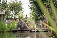 A seated woman wearing a dark grey bathrobe and a man in swimming trunks standing on a jetty on the bank of a wellness pond, surrounded by grass and trees.