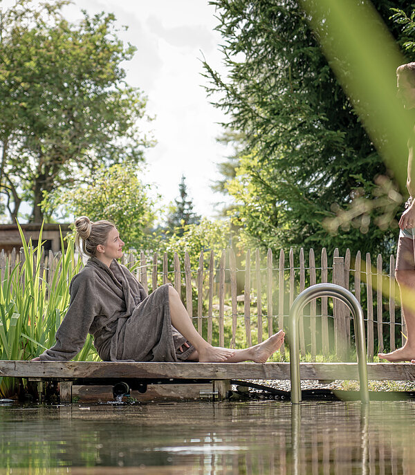 Une femme vêtue d’un peignoir gris foncé est assise, tandis qu’un homme en maillot de bain se tient debout sur un ponton au bord d’un étang dédié au bien-être, au milieu de l’herbe et des arbres.