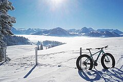 Im Vordergrund ist ein Wintermountainbike in verschneiter Winterlandschaft zusehen, dahinter ist ein Nebelmeer mit einem Bergpanorama zu sehen.