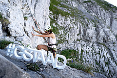 Une femme grimpe sur un rocher dans une via ferrata, au premier plan un petit logo blanc #Gstaad.
