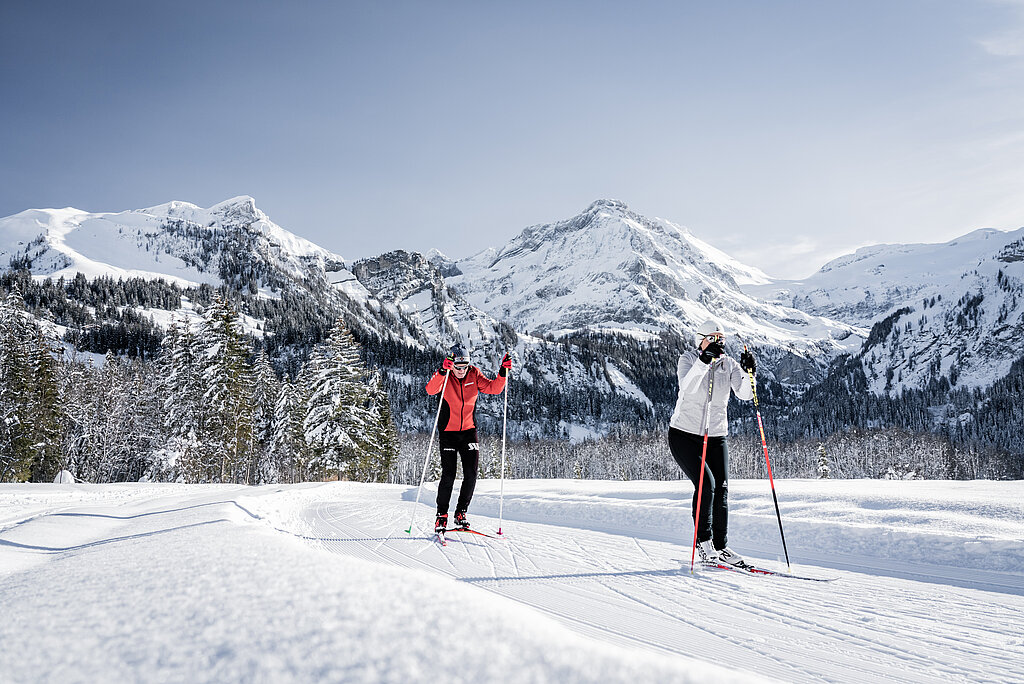 <p>Ein Ehepaar, in rot und weiss gekleidet, am Langlaufen auf einer Loipe vor verschneitem Bergpanorama.</p>