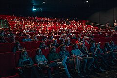 Kinosaal mit roten Sesseln und vielen Menschen die auf die Leinwand schauen.