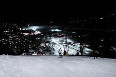 Skieurs et snowboarders sur une piste de ski éclairée, surmontée d'une télécabine rouge. En arrière-plan, un village éclairé avec une gare.