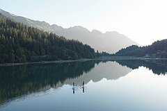 Zwei Personen beim Stand-Up-Paddling auf einem stillen Bergsee, umgeben von Wäldern und Bergen, Spiegelung im klaren Wasser.